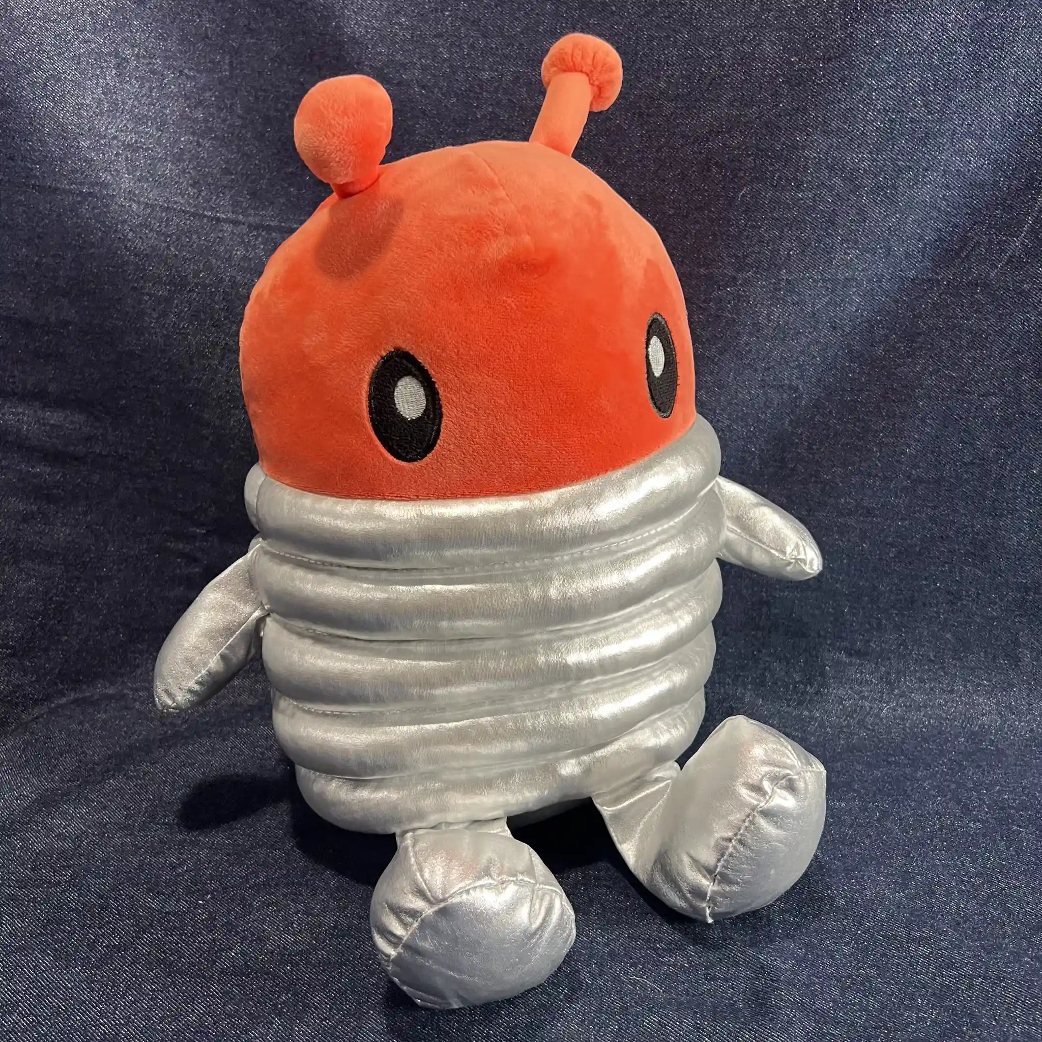 A plush toy character with a bright orange-red round head and antennae, black-and-white eyes, and a ribbed metallic silver body, sitting against a dark blue fabric backdrop.