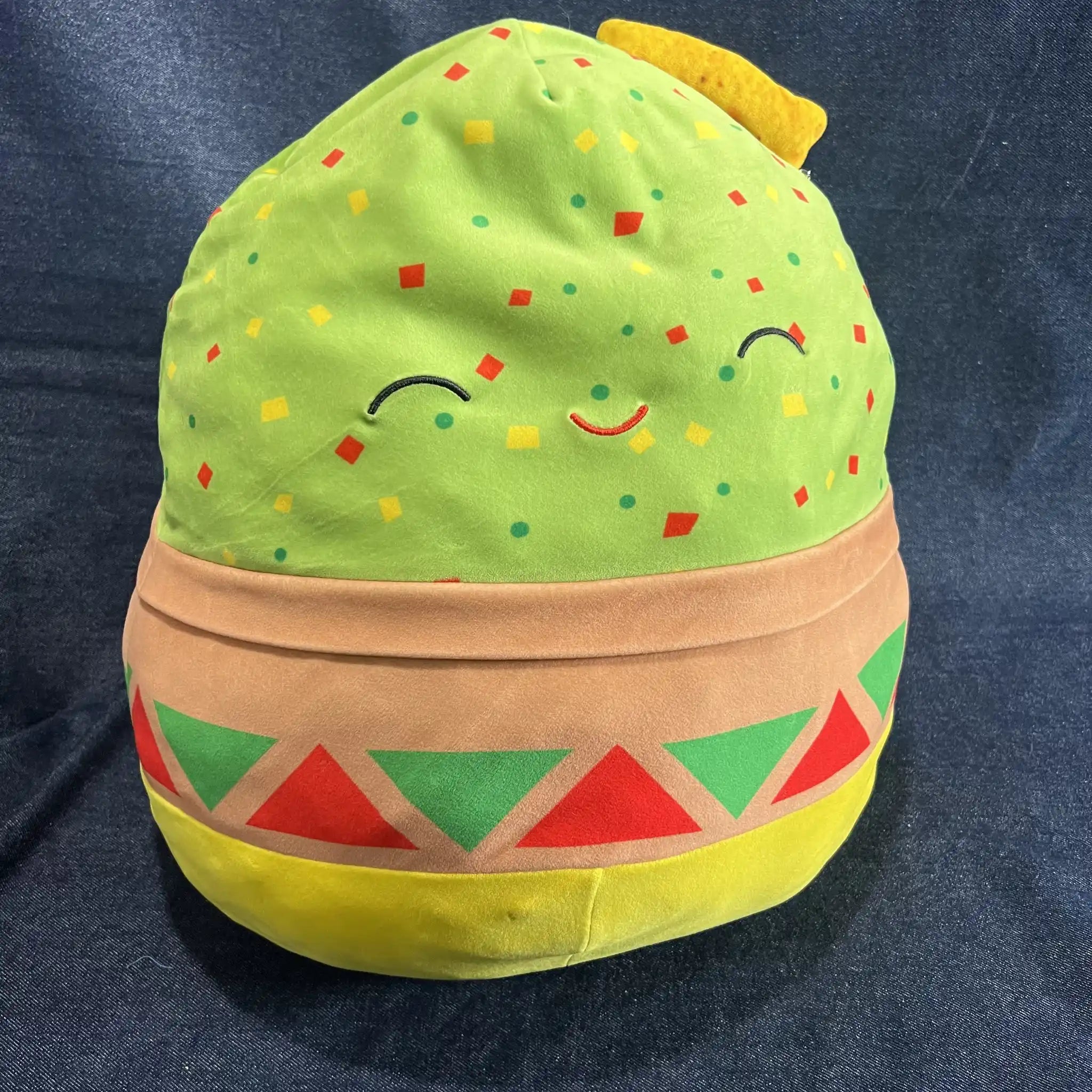 A plush toy shaped like a smiling bowl of guacamole, set against a dark blue fabric background.