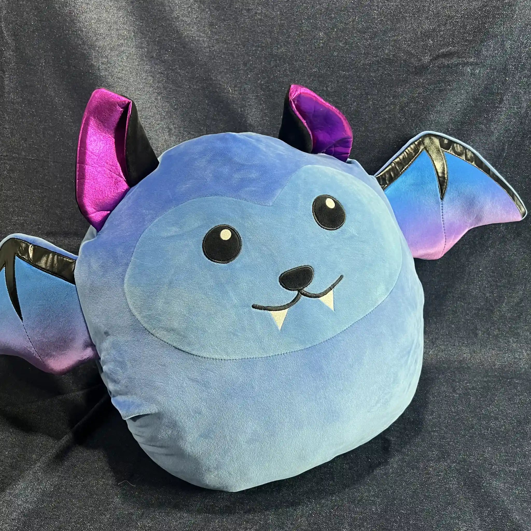 A plush bat-shaped pillow in light blue with purple and iridescent blue wings, black embroidered eyes and nose, and small white fangs, photographed against a dark gray fabric backdrop.