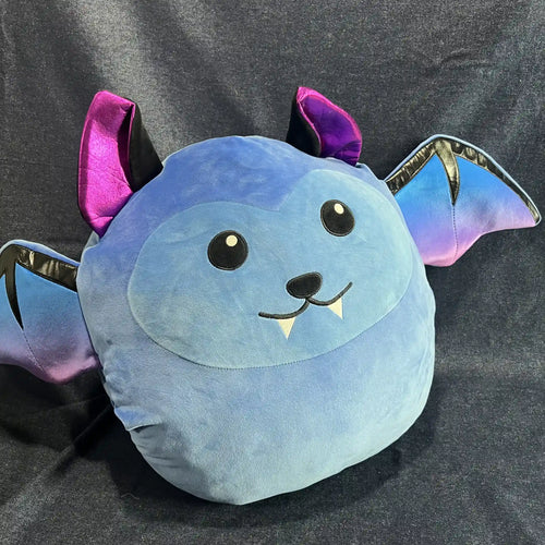 A plush bat-shaped pillow in light blue with purple and iridescent blue wings, black embroidered eyes and nose, and small white fangs, photographed against a dark gray fabric backdrop.