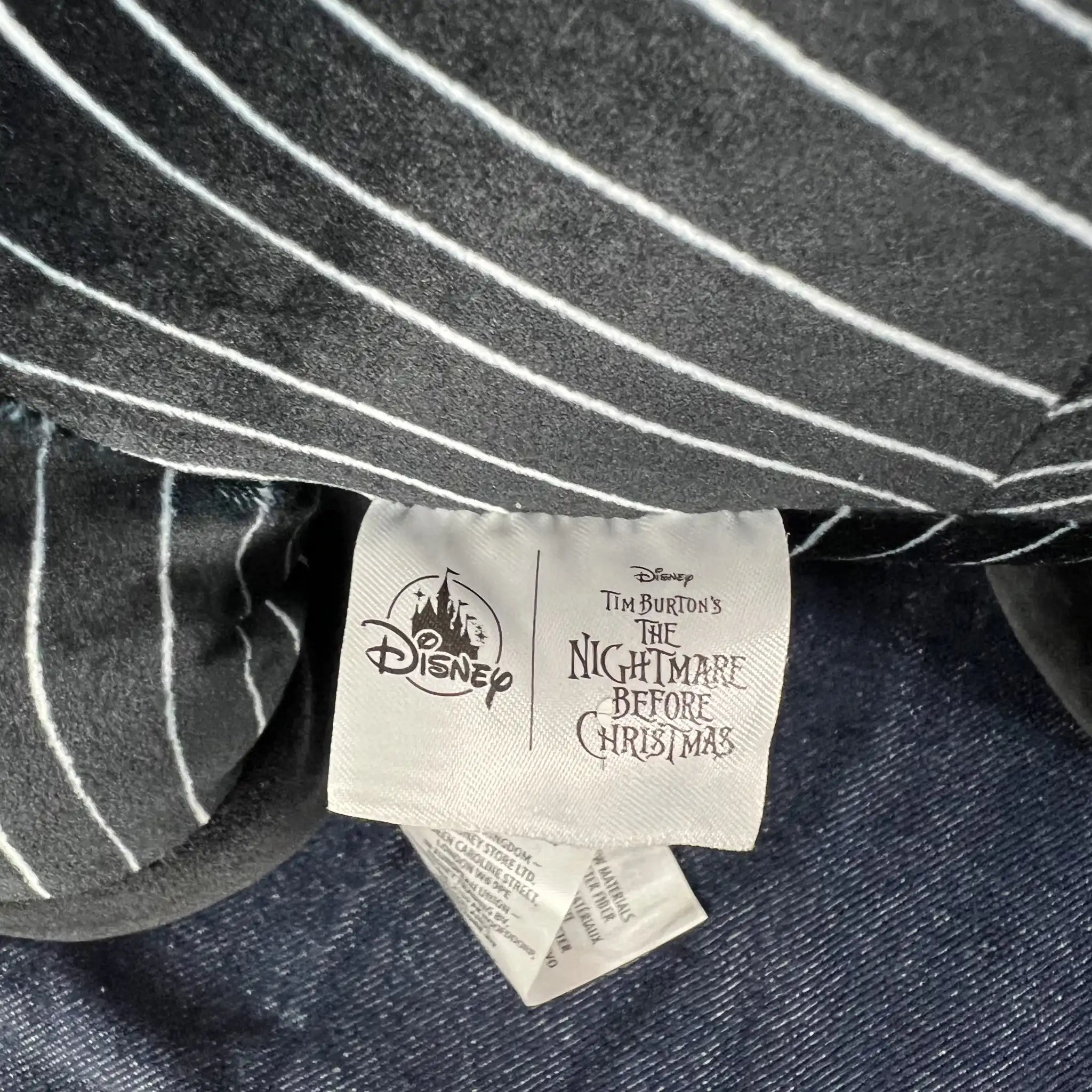 Close-up of a Disney Tim Burton's The Nightmare Before Christmas garment label sewn onto a black and white pinstriped fabric item, with a dark blue denim-like fabric visible underneath.